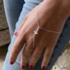 Bijou gothique élégant porté sur une main qui pend le long d'un pantalon : bracelet bague avec croix en argent et chaîne pour main, signé Ninanina