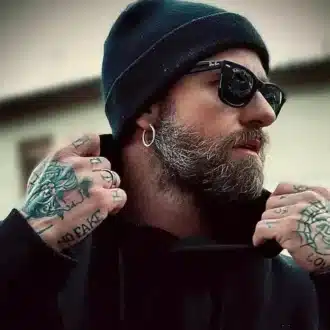 Boucle d’oreille homme en acier inoxydable, créole minimaliste de 20 mm pour un style épuré portée par un homme barbu et tatoué avec lunettes et bonnet noir