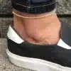 Un bracelet de cheville élégant en perles blanches naturelles, conçu pour hommes et femmes, présenté sur une cheville vêtue d'un jean et de chaussures décontractées en toile noire et blanche. Le design minimaliste de ce bijou s'adapte parfaitement à un style casual tout en ajoutant une touche raffinée.