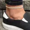 Un bracelet de cheville élégant en perles blanches naturelles, conçu pour hommes et femmes, présenté sur une cheville vêtue d'un jean et de chaussures décontractées en toile noire et blanche. Le design minimaliste de ce bijou s'adapte parfaitement à un style casual tout en ajoutant une touche raffinée.